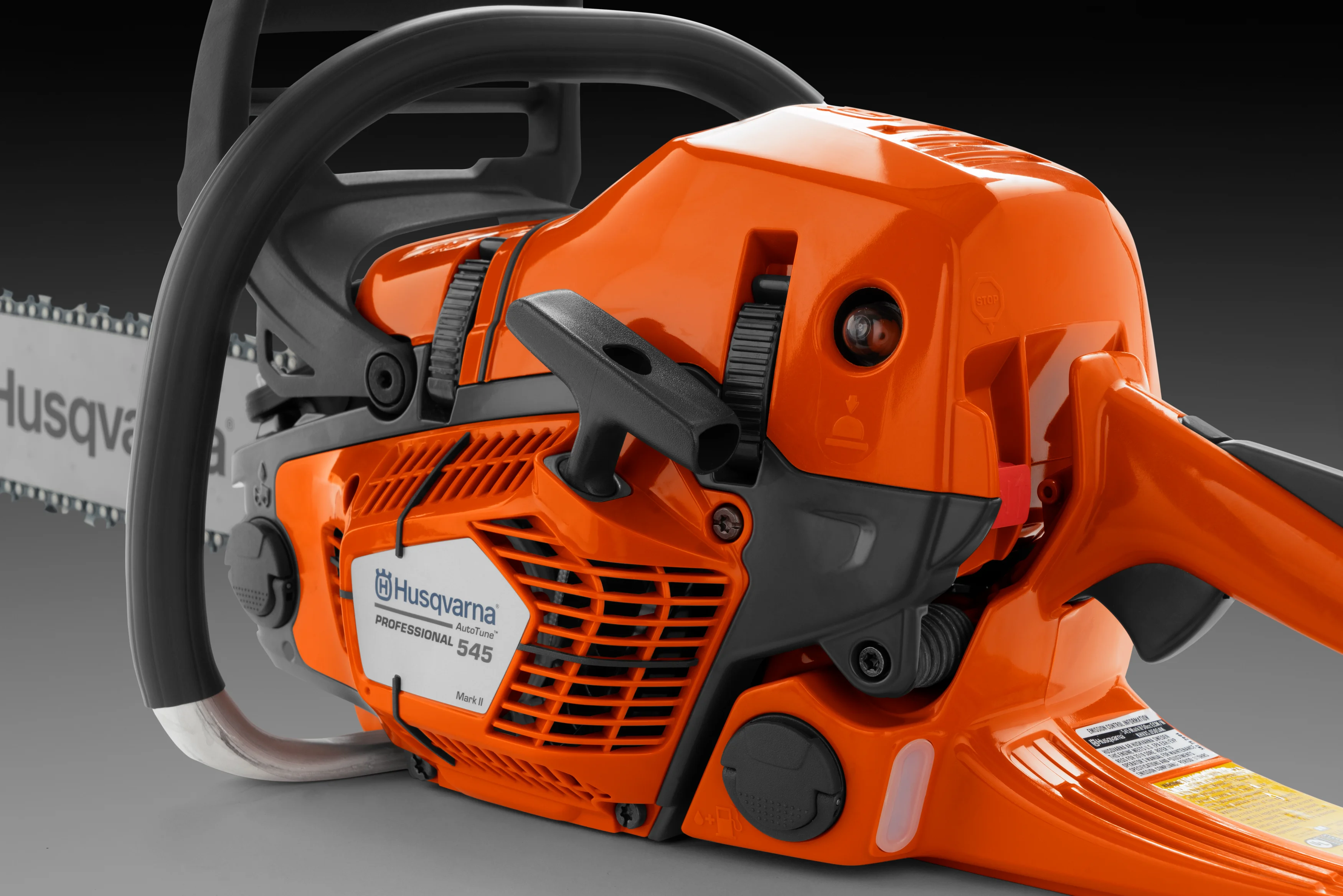 Nahaufnahme der linken Seite einer Husqvarna 545 Mark II Motorsäge auf grauem Hintergrund. Gut sichtbar sind der Startergriff, die Kraftstoffpumpe, der Typenaufkleber und der graue Griffbügel.