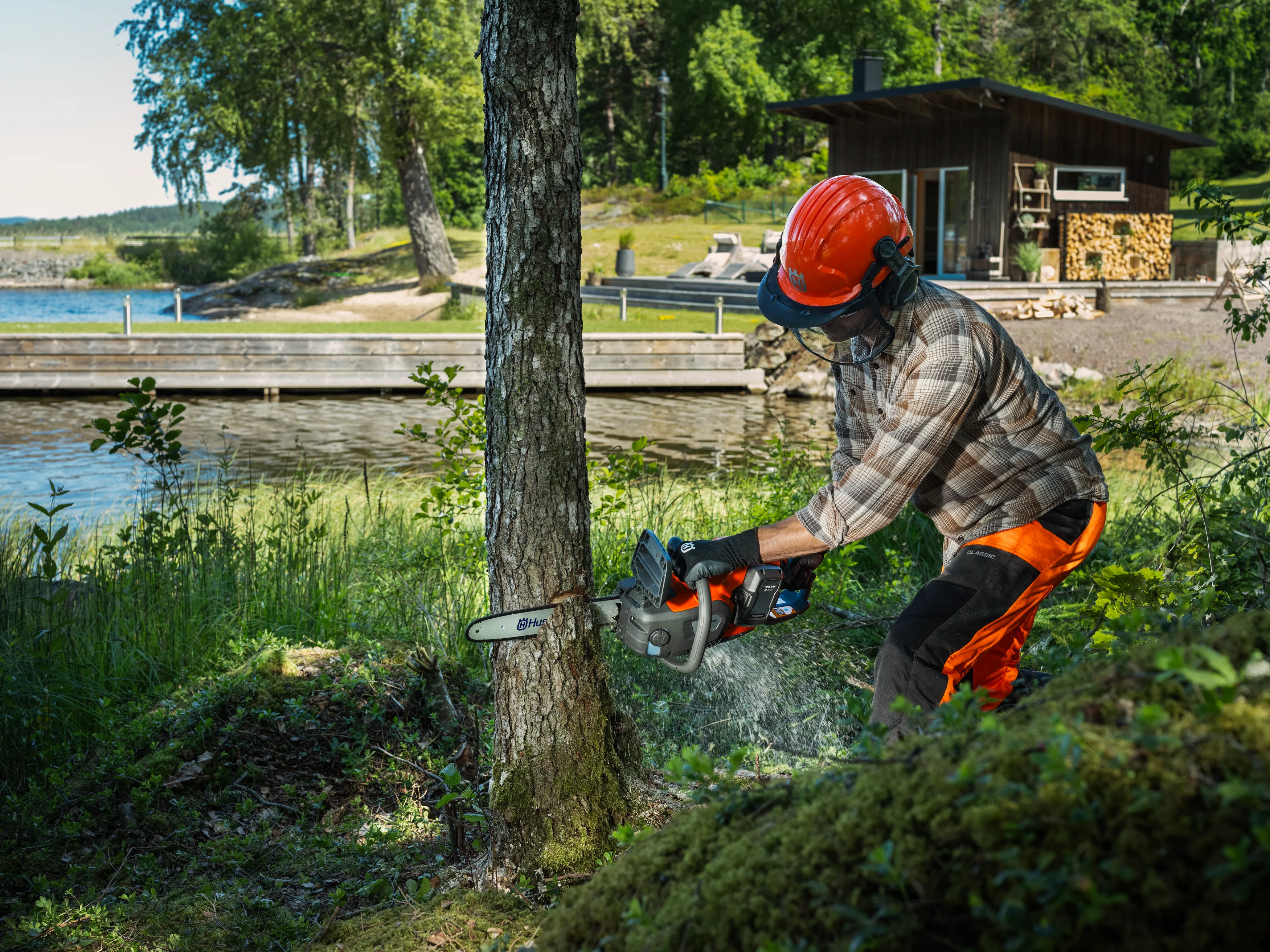 Forstarbeiter mit Helm, Gehörschutz und Schnittschutzbekleidung fällt einen dünnen Baumstamm mit der Husqvarna 242i Akku-Motorsäge am Seeufer.