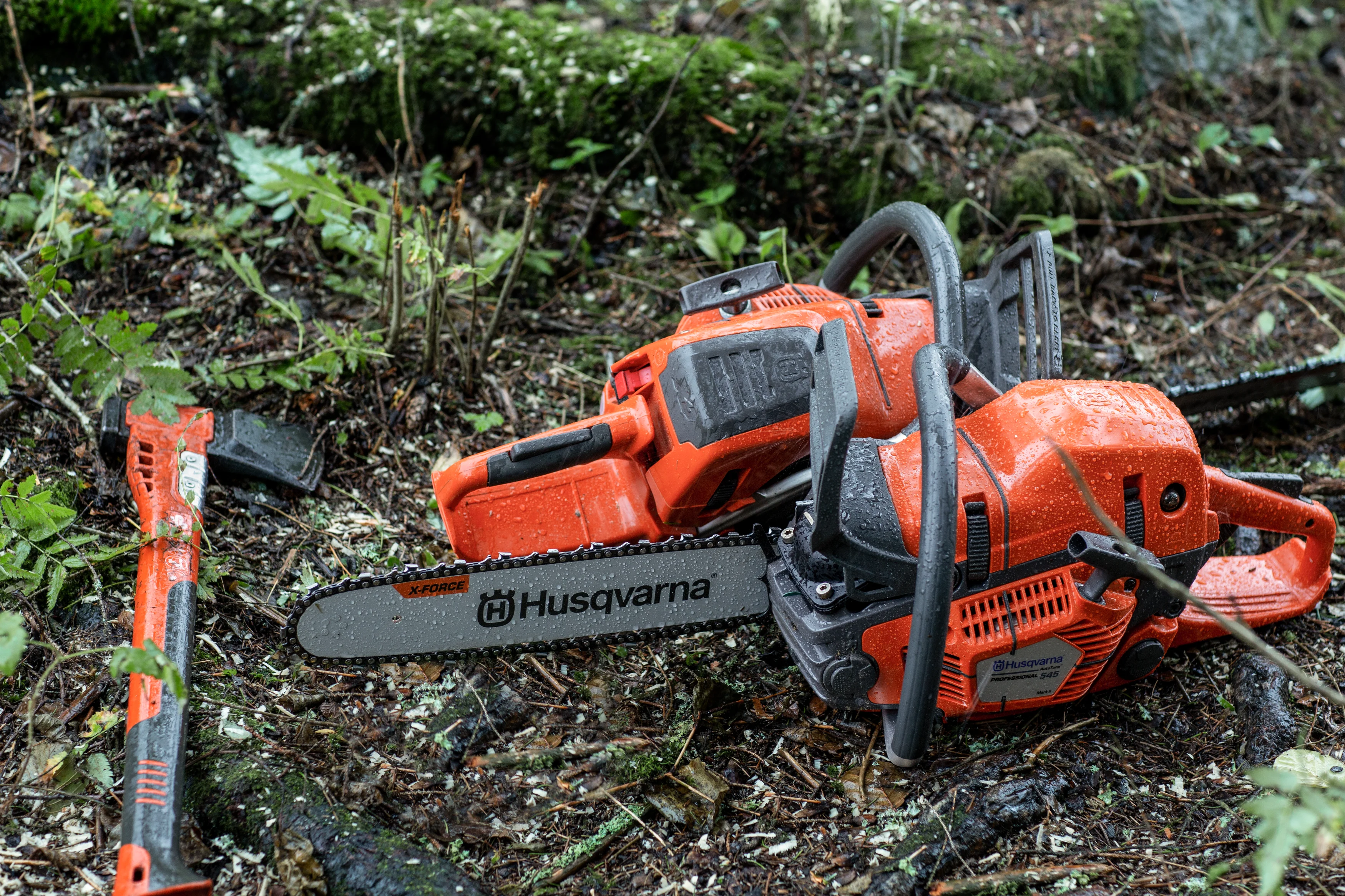 Zwei nasse, orangefarbene Husqvarna Motorsägen liegen zusammen mit einem Beil mit orangefarbenem Stiel auf feuchtem Waldboden.