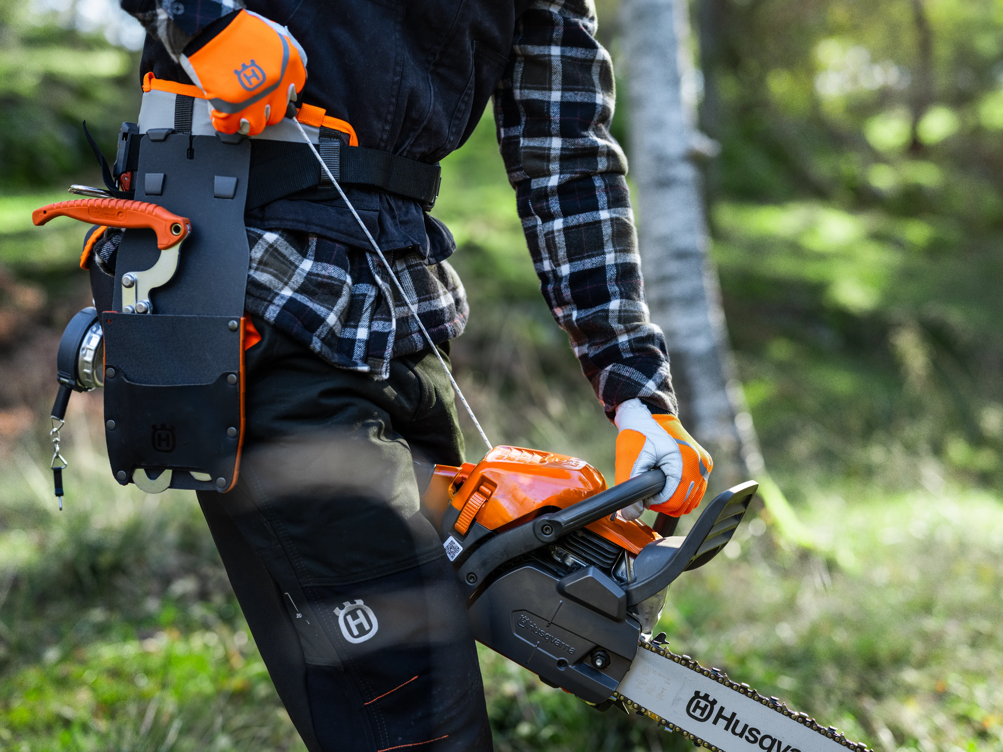 Person in Schutzkleidung trägt eine Husqvarna Motorsäge am Griff. Ein Werkzeuggürtel mit Holster und einem Werkzeug mit orangefarbenem Griff ist an der Hüfte sichtbar.