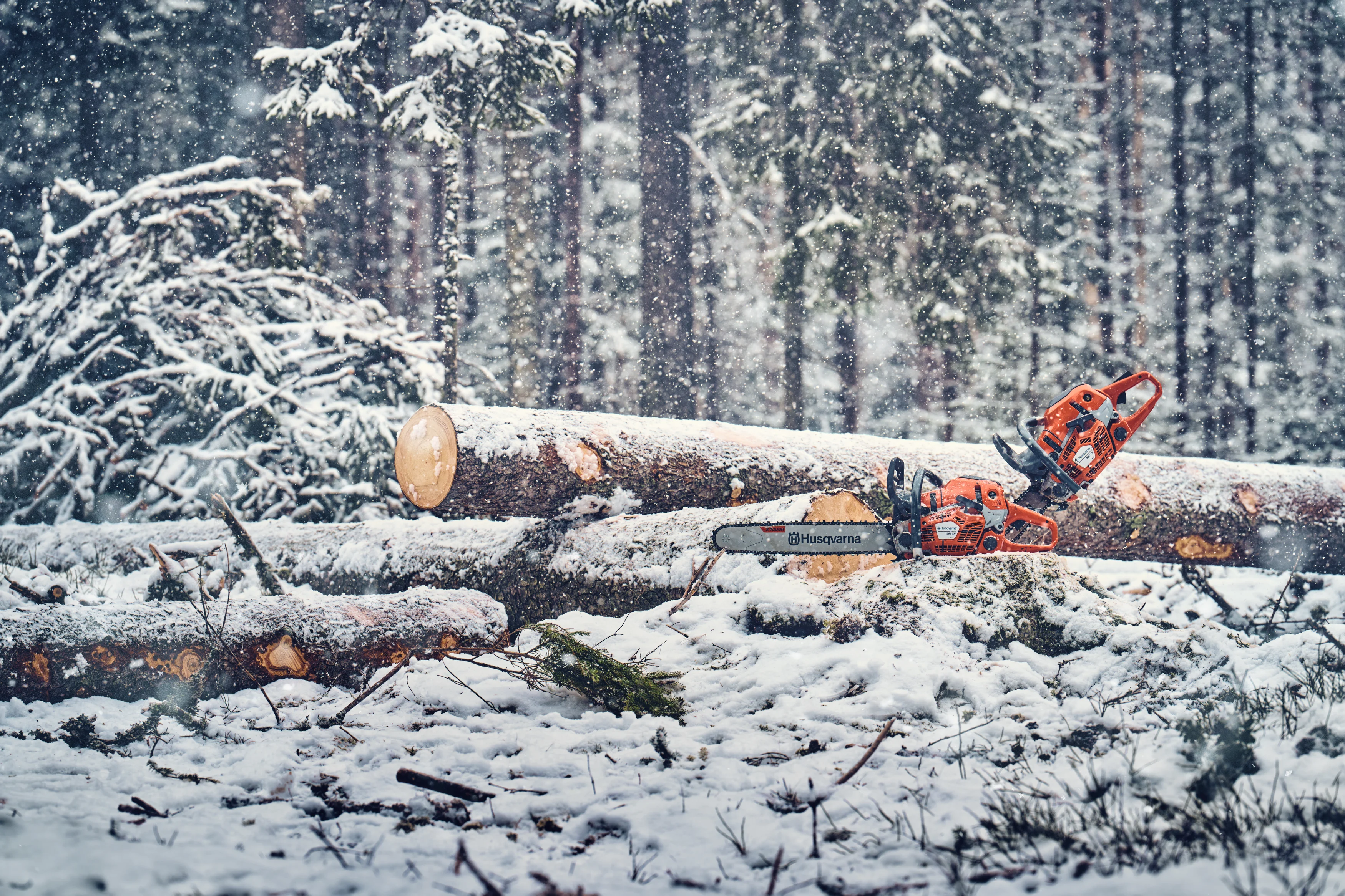 Zwei orangefarbene Husqvarna Motorsägen liegen auf schneebedeckten Baumstämmen in einer Winterlandschaft.