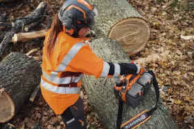 Forstarbeiterin in Warnkleidung und Schutzhelm greift nach einer Husqvarna Akku-Tasche mit Akkus, die auf einem Baumstamm im Wald steht.