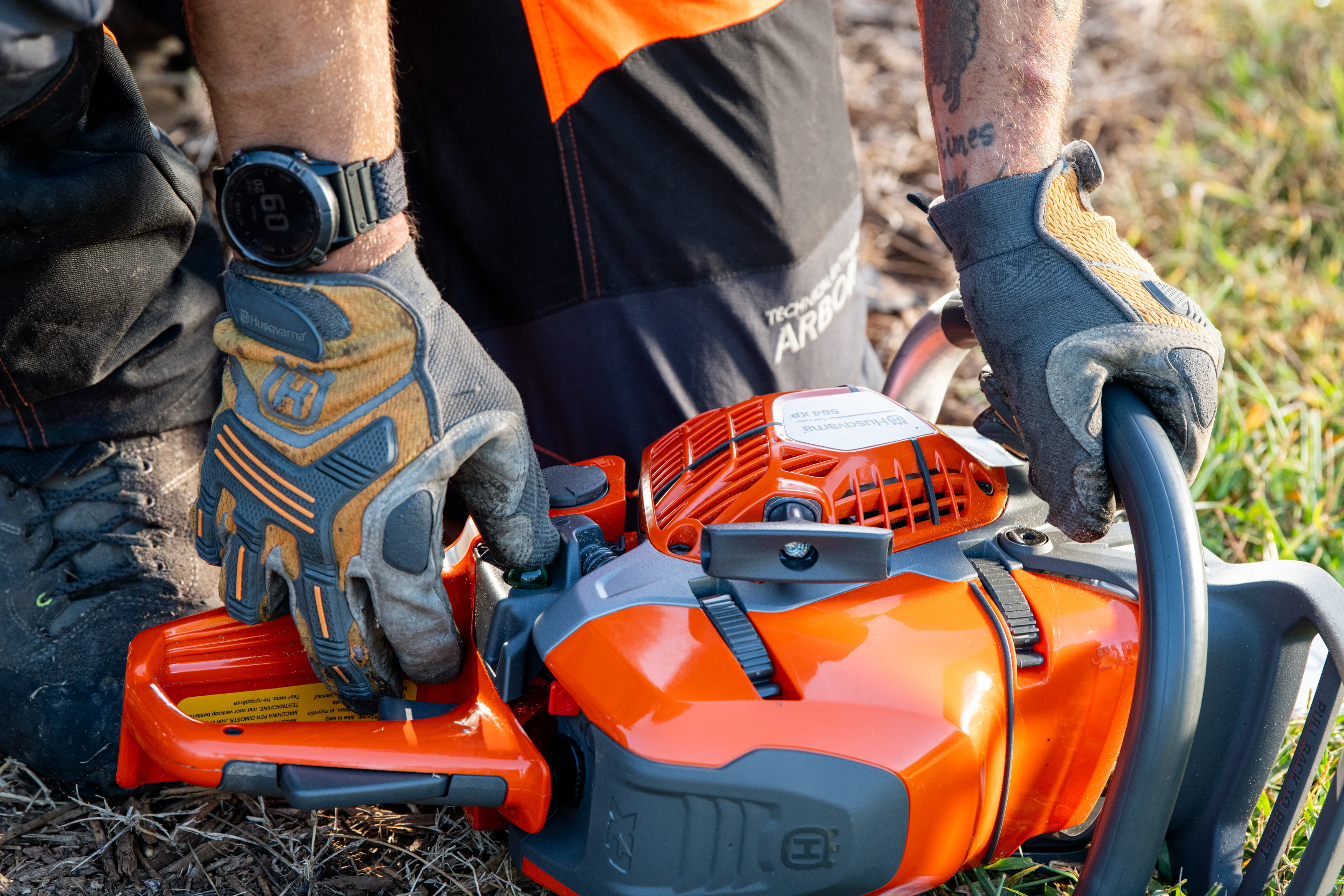 Nahaufnahme der Bedienelemente und der Startvorbereitung einer Husqvarna Motorsäge mit Arbeitshandschuhen.